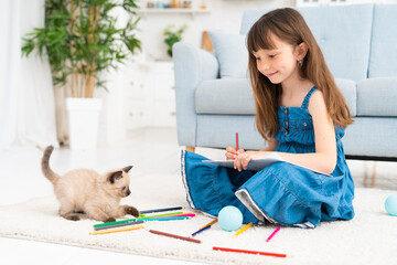Child girl painting with kitten and sitting on the floor. Little female person drawing and playful kitten next to her at home playing with colorful pencils.