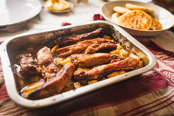 grilled pork ribs on table inside oven aluminum container with venezuelan arepas and potatoes, table cloth, and warm natural light