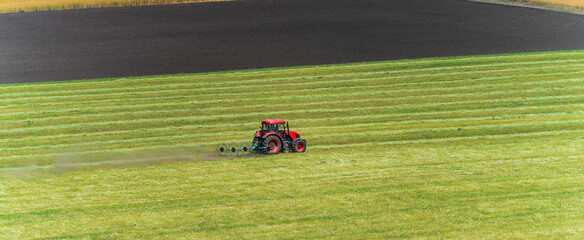 Red tractor mowing on green cultivated agriculture green field, panoramic image.
