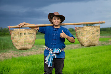 Handsome Asian man farmer carries baskets on shoulders to work at paddy field, wears hat, blue shirt,  tied loincloth on waist. Dark sky before raining background. Concept : Agriculture occupation.
