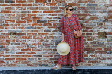 A stylish aged woman in a folklore dress and sunglasses against a brick wall. Summer fashion for older women. - 516951984