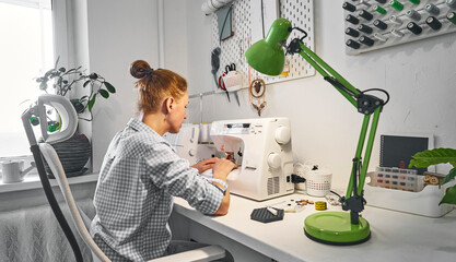 A seamstress girl at work in a comfortable home workshop with overlock equipment and a sewing machine.