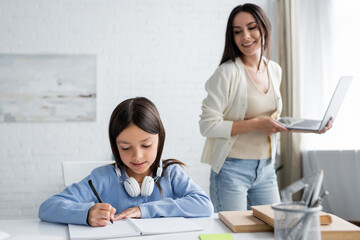 smiling babysitter with laptop near girl writing in notebook at home.