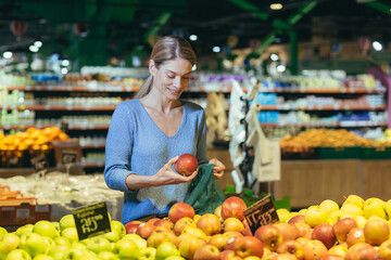A woman in a supermarket, a buyer chooses an apple fruit, buys and puts an ecological bag in the basket