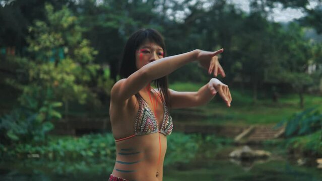 Elegant Asian girl with painted body performing an extatic dance in a lush tropical forest near calm clean pond. 