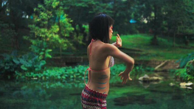Elegant Asian girl with painted body performing an extatic dance in a lush tropical forest near calm clean pond. 