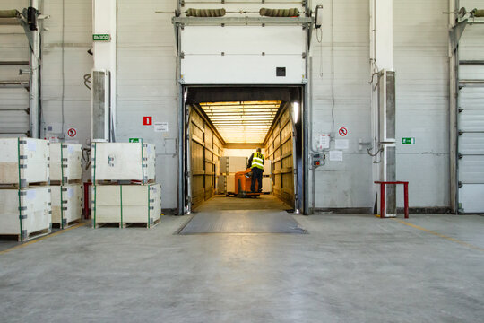 Interior of a modern warehouse storage of retail shop with pallet truck near shelves and loading ramps. Storehouse worker in uniform working on forklift.