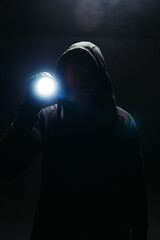 Silhouette of bandit in hoodie holding flashlight on black background with smoke