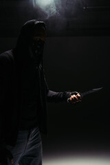 Silhouette of african american robber in mask and hoodie holding knife on black background with...