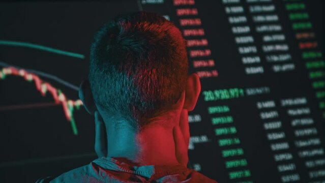 Trader analyze cryptocurrency charts on a big screen in the dark. Rear view of man in red-blue lighting looking at currency charts in stock market. Trading stocks, bitcoins, futures, investment market