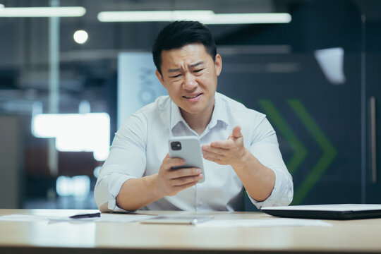 Shocked And Upset Young Man Asian Businessman Playing Games On Mobile Phone For Money At Work,lost,received Bad News By Message. Sitting Sad At The Desk In A Modern Office.