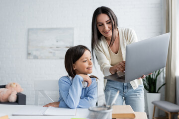 Obraz na płótnie Canvas babysitter showing laptop to smiling girl doing homework on blurred foreground.