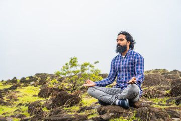 Naklejka na ściany i meble Young beard man with eyes closed doing meditation or yoga on top of hill - concept of self caring, health care and wellness