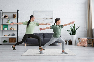 smiling child and babysitter training in warrior pose in modern living room.
