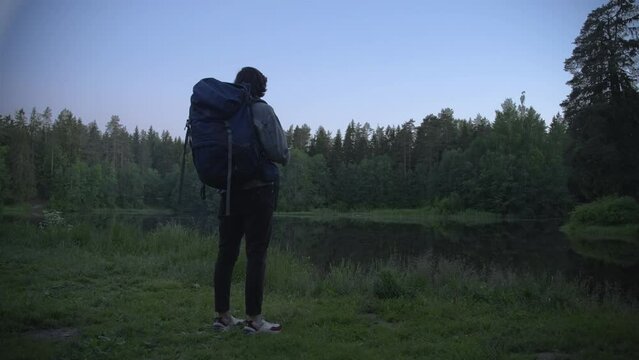A tourist with a backpack approaches a forest lake, stops, takes a smartphone out of his pocket, takes pictures. Summer night. Back view. Slow motion 4k footage