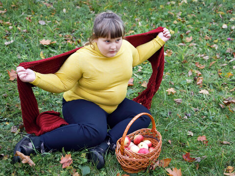 Obese girl with basket of apples sits on grass in autumn and wrapped in scarf