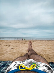 A man with hairy legs lies on the beach throwing his leg over his leg. Overcast skies on the beach. Swimming trunks with palm trees.