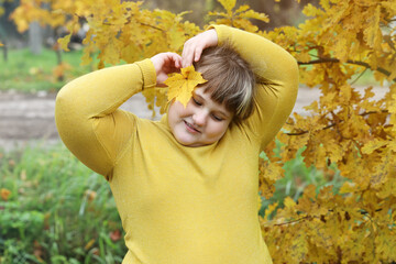Obese smiling girl stands outdoors and attaching yellow maple leaf for right ear