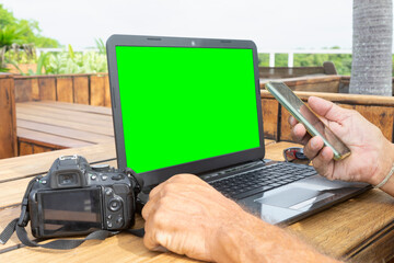 Close-up of a freelance photographer working with a smartphone, laptop with a green screen empty SLR camera on an outdoor wooden table. 