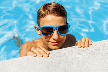 cute boy in sunglasses leaning on the edge of the pool during summer vacation. The boy looks at the...