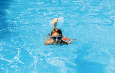 Boy in sunglasses swims in the pool, smiling and having a happy day. Cute happy little boy swims and dives in the pool