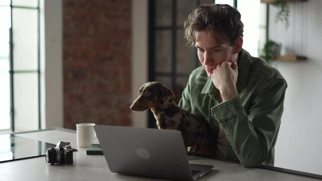 4k Young man is using laptop and sitting at table with dachshund dog at home spbd. Close view of handsome caucasian man looks at computer screen with smile and does work, spends time with cute pet and