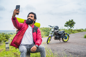 Happy smiling beard man making video call on mobile phone in front of motorbike while sitting on roadside - concept of technology, social media sharing and travel vloggs.