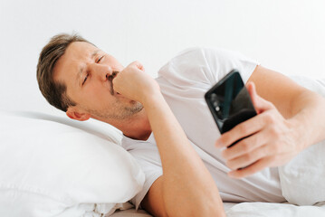 Sleepy yawning young man holding mobile phone and looking at alarm clock while lying in bed in early morning. Wake up tired guy using smart phone in bedroom indoors
