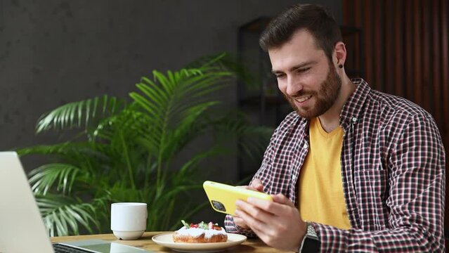 Young man 20s wear casual clothes sits alone at table in coffee shop cafe work on laptop pc use cell phone doing winner gesture relax rest in restaurant during free time indoor. Mobile office concept
