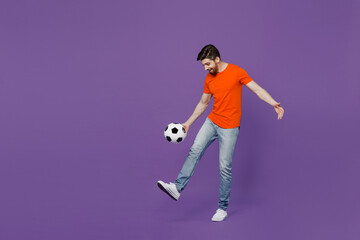 Fototapeta na wymiar Full size young cheerful excited fun fan man he 20s wear orange t-shirt cheer up support football sport team juggling soccer ball on leg watch tv live stream isolated on plain dark purple background.