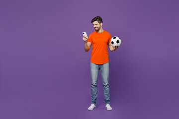 Full body young fun fan man he wear orange t-shirt cheer up support football sport team hold in hand use mobile cell phone soccer ball watch tv live stream isolated on plain dark purple background