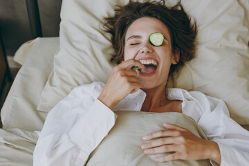 Young fun woman wear white shirt pajama she lying in bed with mask on eyes eat biting cucumber...