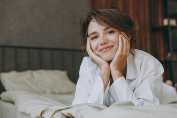 Young smiling fun woman wear white shirt pajama she lying in bed look camera prop up chin rest...