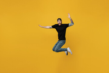 Fototapeta na wymiar Full body young excited satisfied jubilant bearded tattooed man 20s he wears casual black t-shirt cap jump high with outstretched hands isolated on plain yellow wall background. Tattoo translate fun.