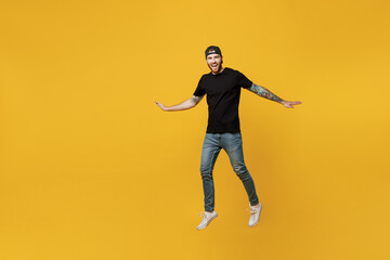 Fototapeta na wymiar Full body young smiling bearded tattooed man 20s he wears casual black t-shirt cap jump high with outstretched hands isolated on plain yellow wall background studio portrait. People lifestyle concept.