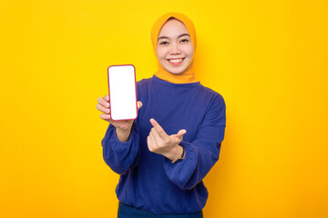 Excited young Asian Muslim woman dressed in casual sweater showing blank screen mobile phone, recommending mobile app isolated over yellow background