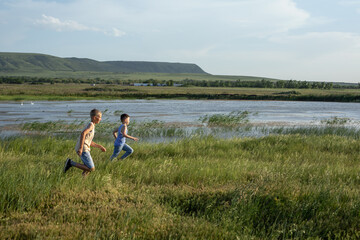 Two brothers in jeans and t-shirts run along the pond competing in speed. Summer sunny day near the...