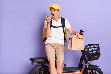 Portrait of angry courier man wearing white t shirt and cap delivering parcels on electric bike isolated over purple background, using voice assistant or recording message, talking with client.