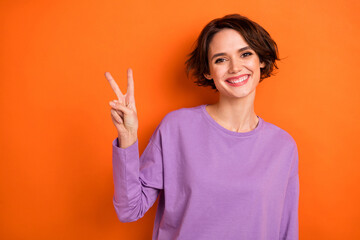 Photo of cheerful adorable girl toothy smile hand fingers demonstrate v-sign isolated on orange...