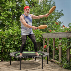 senior man is exercising on a mini trampoline with wooden Indian clubs in a backyard patio, fitness...