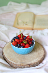 Bowl of strawberries, cherries and blueberries, open book and reading glasses on the bed. Selective focus.