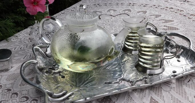 Female hands picking up a cup with mint tea on a metal tray, a teapot with green tea, a table with a vase of pink flowers, a thin brown knitted tablecloth. Tea time concept in the garden