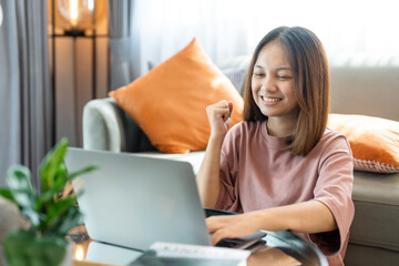 A young Asian woman with long hair sitting in the living room on the sofa using a laptop to video call a coworker. showing a happy expression. Work from home.