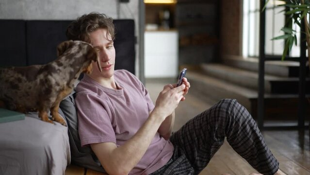 4k Young man is using smartphone and sitting on floor with dachshund dog at home spbd. Close view of caucasian guy holds device in his hand and looks with smile, plays with cute pet and sits in
