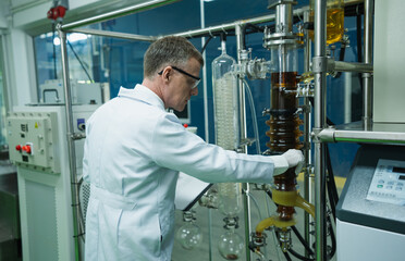 Portrait of Caucasian senior man researcher with lab coat, gloves and glasses adjust cannabis extraction machine. Expert male biochemical control process of quality checking CBD oil from marijuana.