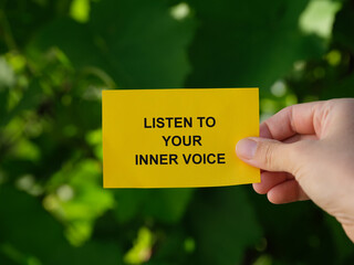 A woman holding a yellow paper note with the words Listen To Your Inner Voice  on it against a background of foliage.