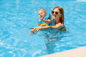 Fototapeta na wymiar Happy young mother plays with her baby in the outdoor pool on a hot summer day. Children learn to swim on a family vacation. A young family with a child has fun in the pool on a summer day