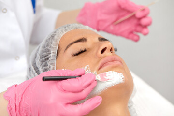 Beautician uses brush to apply white mask made of kaolin clay on half of woman's face. Close up of young woman in white headband lying on soft towel in beauty salon. Concept of cosmetic care for face
