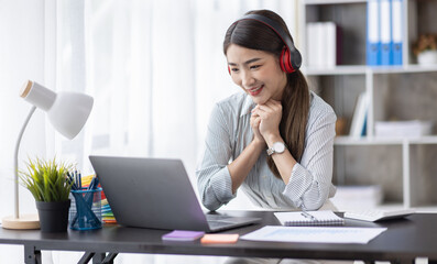 Young Asian woman sitting at desk and connecting with her laptop, she is wearing headphones and working