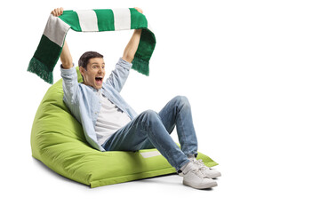 Young man cheering with a green and white scarf and sitting on a bean bag armchair
